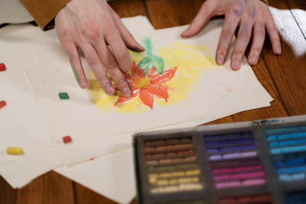 Mains posées sur une feuille autour d’une fleur dessinée aux pastels secs, boîte de pastels au premier plan