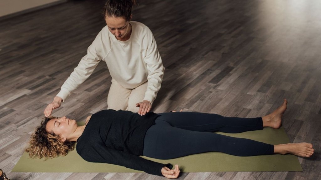 Femme allongée et habillée sur un tapis, les yeux fermés, pendant qu’une praticienne place ses mains près de sa tête